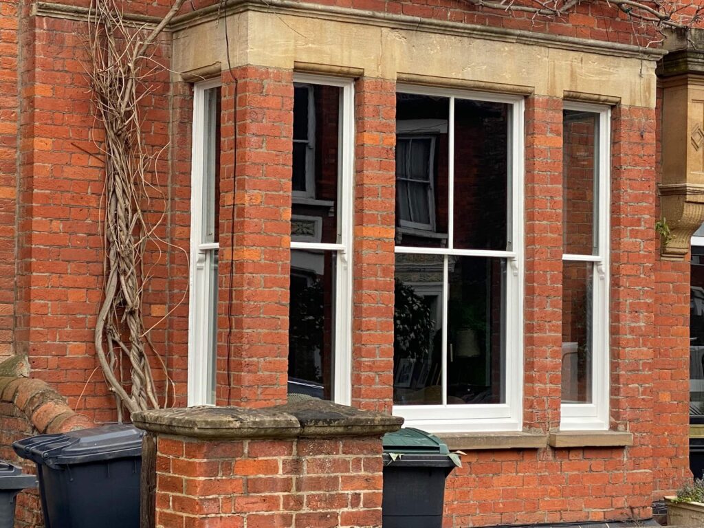 A brick building with a large bay window featuring white frames, two rubbish bins, and a bare climbing plant on the wall—ideal for those interested in sash window repair South London.