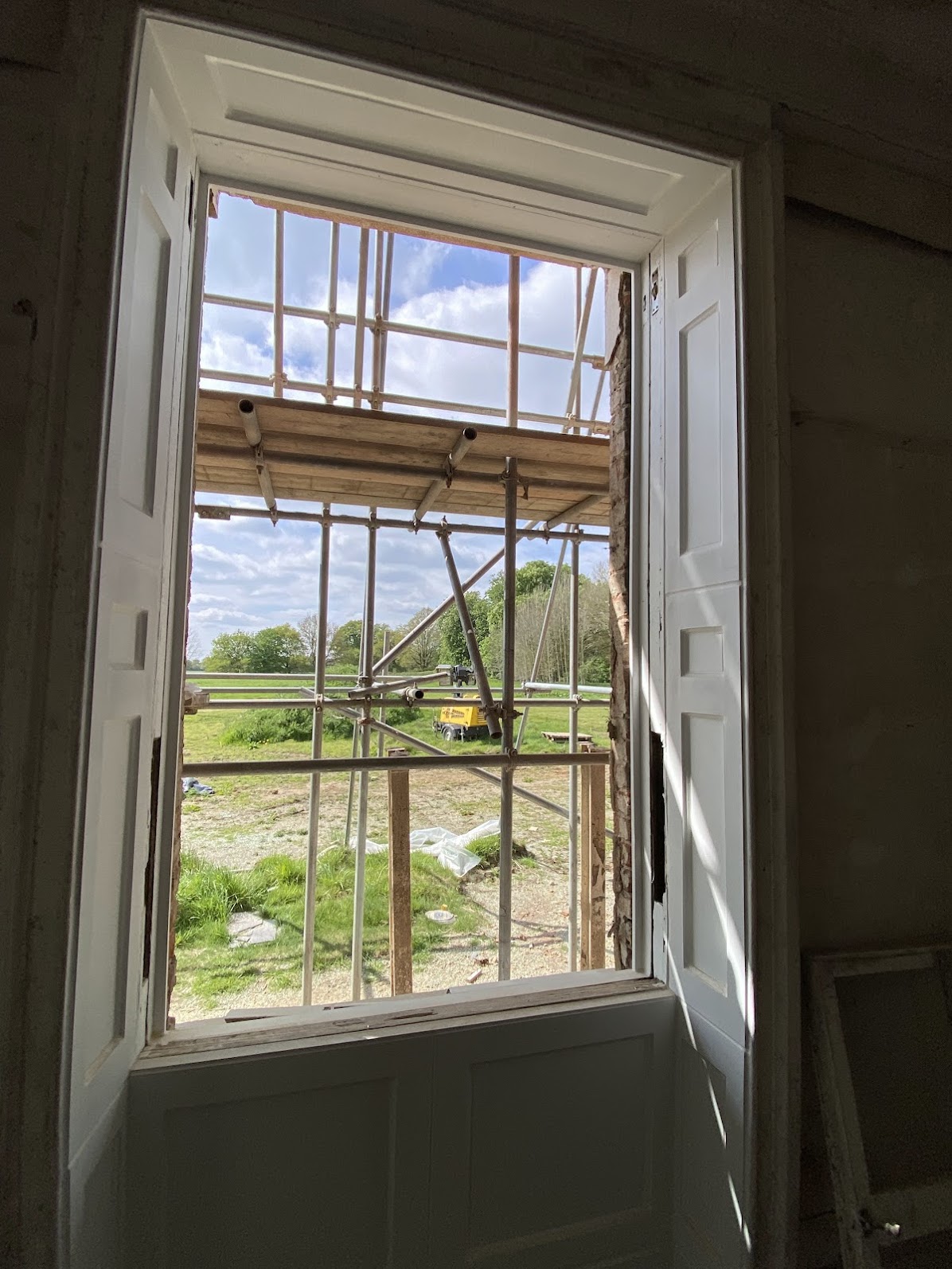 Open window with white shutters showing scaffolding outside and a grassy area with trees and a yellow object in the distance under a partly cloudy sky, ideal for those seeking sash window repair South London services.