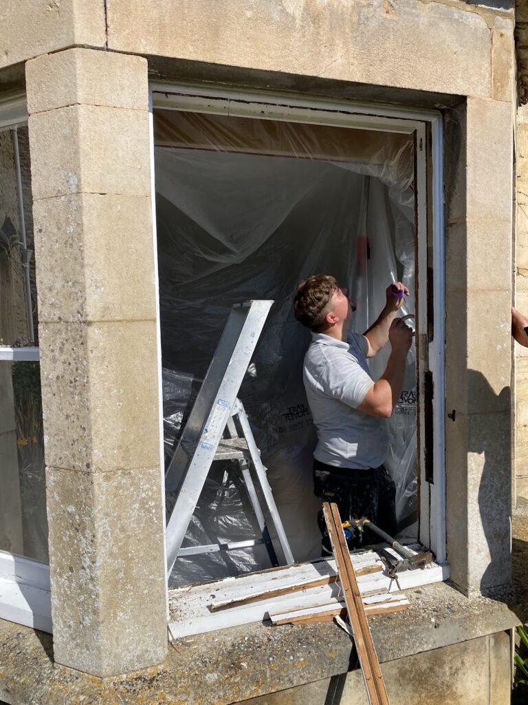 A person stands in a window frame, painting or repairing its edges. Nearby, a ladder, plastic sheet, and wooden tools suggest Sash Window Repair South London expertise at work.