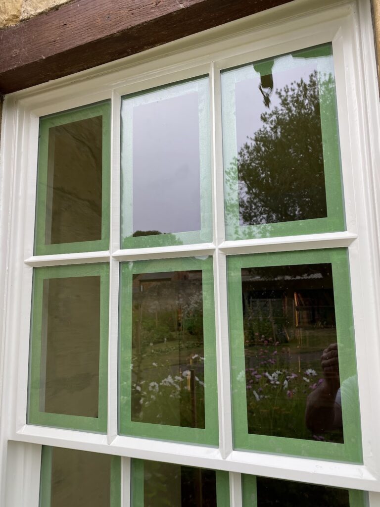 A multi-pane window with green masking tape around each pane, reflecting an outdoor garden scene—an ideal example for those seeking Sash Window Repair South London.
