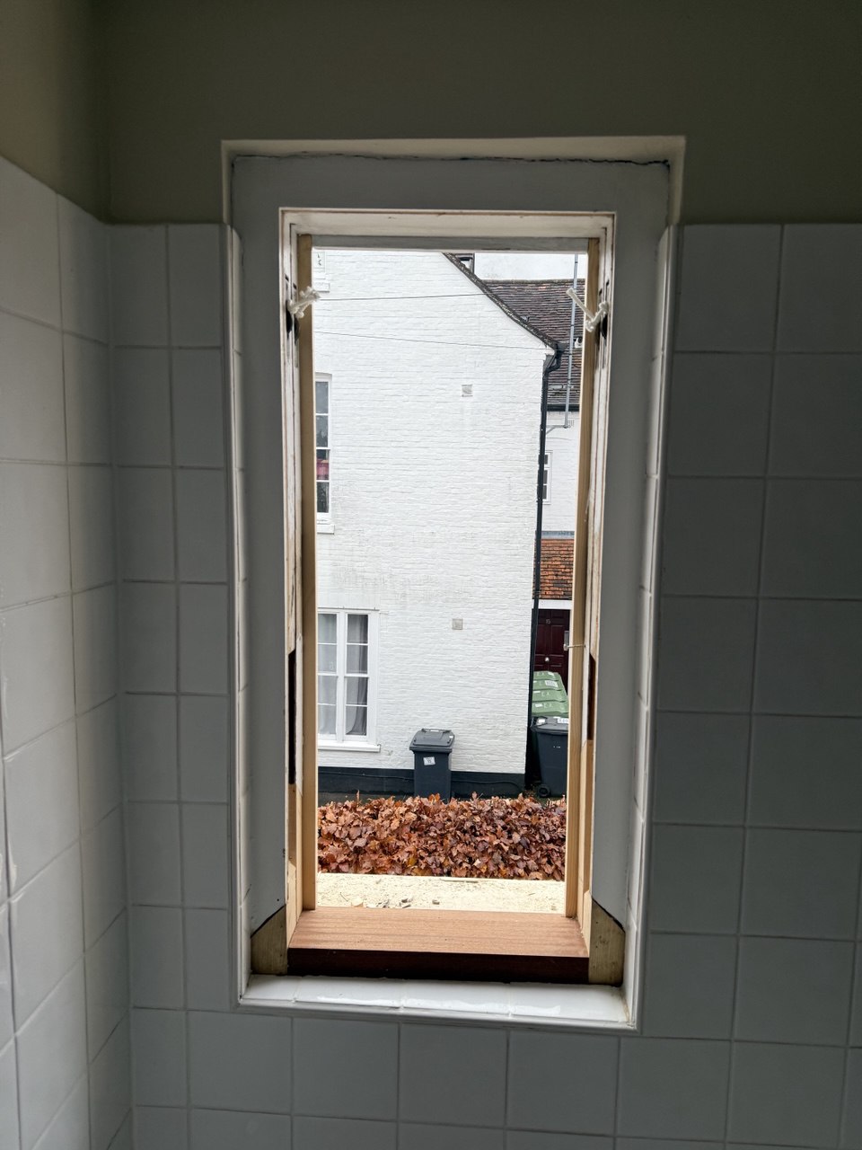 Open rectangular window in a tiled wall, showing a view of a white building, rubbish bins, and fallen brown leaves outside—perfect inspiration for Sash Window Repair South London.
