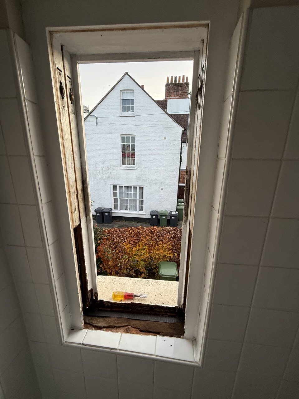 View through a narrow, partially damaged window highlights a white house, several rubbish bins, and autumn foliage outside—perfect inspiration for those seeking sash window repair South London solutions.